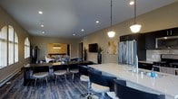 This single-floor space offers a well-designed, open-plan layout that seamlessly integrates multiple functional zones within one cohesive environment. The kitchen area is outfitted with stainless steel appliances, including a full-size refrigerator, and features a generous light-toned countertop island with bar-height seating — ideal for casual gathering or meal preparation. Pendant lighting with warm globe fixtures hangs above, adding a polished accent to the space.

Adjacent to the kitchen, a sizeable dining area accommodates a long dark-wood table surrounded by contemporary chair seating, offering ample room for group meals or collaborative use. The flooring throughout features a rich, multi-toned wood-plank design that adds depth and visual interest underfoot.

Recessed lighting across the ceiling provides consistent, even illumination throughout the space, while neutral wall tones create a clean and inviting backdrop. Decorative potted plants are strategically placed to introduce natural elements and warmth.

Further into the layout, a secondary lounge or recreation area is visible, separated by a partial wall and featuring additional seating and ambient lighting. A wall-mounted television and colorful artwork add personality to the space, rounding out a versatile, modern environment suited for both social and functional use.