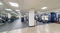 Located at 1001-1003 Easton Rd in Willow Grove, PA, this fitness center offers a generously sized, single-floor exercise facility designed to support a full range of workout activities. The space is divided into distinct functional zones, all unified by a bold, energetic design aesthetic featuring navy blue accent walls adorned with motivational fitness typography in white and orange.

The main workout floor is finished with durable rubber flooring and houses an extensive array of strength training machines and equipment, along with a dedicated row of cardio machines including treadmills, ellipticals, and stationary bikes. Large wall-mounted mirrors amplify the sense of space and allow members to monitor their form. The area is well-lit throughout by recessed panel lighting set into a drop ceiling, creating a bright and consistent environment.

A separate studio area features smooth hardwood-style flooring, making it well-suited for stretching, group exercise, or functional training. This room is lined with the same motivational wall graphics and benefits from natural light through a series of windows overlooking the outdoors.

Additional amenities include a water fountain station set against a vibrant yellow accent wall, along with convenient access doors throughout. The overall design is clean, modern, and purpose-built to create an energizing atmosphere for users of all fitness levels.