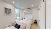 This thoughtfully designed single-floor studio maximizes functionality without sacrificing style. The main living area is bright and airy, finished with light hardwood flooring and crisp white walls that reflect natural light pouring in through multiple large windows framing impressive urban cityscape views. A built-in Murphy bed serves as the centerpiece of the sleeping and living area, allowing the space to adapt seamlessly between rest and relaxation. A comfortable sofa and area rug define the living zone, while a wall-mounted flat-screen TV and white dresser provide practical storage and entertainment. A wall-mounted mini-split air conditioning unit ensures year-round climate comfort. The galley-style kitchenette is efficiently outfitted with white shaker cabinetry, quartz countertops, a subway tile backsplash, stainless steel appliances including a refrigerator, microwave, and dishwasher, and open shelving for added storage. Entry is secured with a keypad-enabled door. The bathroom is elegantly appointed with classic black-and-white hexagonal floor tile, white subway tile walls, a glass-enclosed walk-in shower with black fixtures, a white vanity, and a sliding barn door — a charming design detail that ties the space together cohesively.