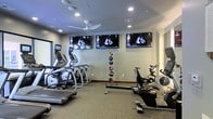 This single-floor fitness center is thoughtfully designed to support a well-rounded cardio and strength training routine. The space features a lineup of elliptical trainers and treadmills along one side, complemented by stationary and air resistance bikes on the opposite side, catering to a variety of cardio preferences. A vertical medicine ball rack stationed at the center adds a functional training element to the room. Three large flat-screen televisions are mounted high along the main wall, allowing exercisers to stay entertained during workouts, while a ceiling fan overhead helps maintain comfortable airflow throughout the space. Recessed lighting illuminates the room evenly, creating a clean and focused atmosphere. The flooring consists of low-pile carpet suited for equipment placement, and the neutral gray walls contribute to a modern, distraction-free environment. Natural light appears to enter through a window or glass panel on one side, adding brightness to the interior. Overall, the space is efficiently arranged to maximize usability while maintaining an open, uncluttered feel.