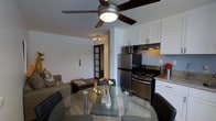 This single-floor space offers a thoughtfully designed open-concept layout that seamlessly connects the living, dining, and kitchen areas into one cohesive environment. The dining area features a round glass-top table paired with sleek modern chairs, creating an elegant yet functional space for meals and gatherings. A ceiling fan with an integrated light fixture overhead provides both comfort and ambient illumination.

The kitchen is well-equipped with stainless steel appliances, including a full-size refrigerator, a gas range with overhead microwave, and a range hood. Crisp white cabinetry extends to the ceiling, maximizing storage, while granite countertops add a refined touch. The clean white palette of the kitchen creates a bright and organized workspace.

The adjoining living area is comfortably furnished with a sofa and a coffee table, providing a relaxed space for lounging. Slatted interior doors add a subtle architectural detail that separates additional spaces beyond, hinting at further rooms within the unit. Light wood-toned flooring runs throughout, tying the spaces together and contributing to the warm, welcoming ambiance. Overall, the layout is efficient and modern, making excellent use of the available space.