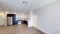 This single-floor space showcases a thoughtfully designed open-concept layout that seamlessly connects the kitchen and living area. The kitchen is a standout feature, equipped with stainless steel appliances including a range, microwave, and dishwasher. Cabinetry combines dark lower cabinets with light upper cabinets, creating a striking two-tone contrast that adds visual depth. A peninsula with a white surround provides a natural transition between the kitchen and living space, offering additional counter space and functionality. Recessed lighting throughout the kitchen ensures ample illumination, complementing the neutral light gray walls. Warm hardwood-style flooring flows continuously across the open area, adding a cohesive and inviting feel to the space. A doorway visible from the main area leads to an additional room, hinting at further functionality beyond the open living zone. The clean, minimalist aesthetic is reinforced by white baseboards, smooth ceilings with a ceiling vent, and an overall uncluttered layout that makes the space feel airy and versatile.