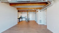 This single-floor unit showcases a compelling blend of industrial and contemporary design elements throughout its open layout. The main living area makes an immediate impression with exposed wood ceiling beams and visible silver ductwork running overhead, adding raw architectural character to the otherwise clean, modern aesthetic. Durable wood-look flooring extends throughout the living spaces, complementing the neutral white walls and recessed lighting.

The kitchen is well-appointed with stainless steel appliances, including a refrigerator, range, microwave, and dishwasher, paired with crisp white shaker-style cabinetry and dark countertops. Sliding barn doors provide a stylish and functional room division within the open-concept living space.

The bedroom offers a quieter atmosphere with soft carpeting underfoot and a window that brings in natural light. Two bathrooms serve the unit: one features a walk-in shower with built-in shelving and a curved curtain rod, while the other includes a full soaking tub and shower combination alongside a white vanity with ample cabinetry. Both bathrooms feature wood-look tile flooring, maintaining a cohesive design throughout the space.
