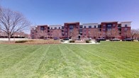 This multi-story residential building presents a classic and distinguished architectural design, constructed with rich red brick and accented with light-colored trim and large windows that line each floor. The exterior is well-proportioned, with multiple balconies offering residents private outdoor space and views of the surrounding grounds.

The building is set within a thoughtfully landscaped campus-style environment. A generous, well-manicured lawn stretches out in front of the structure, creating a sense of openness and providing ample green space. At the heart of the grounds, a central courtyard features a prominent statue as a focal point, surrounded by a circular path, mature trees, and park benches — offering a welcoming area for outdoor relaxation and community gathering.

The grounds are neatly maintained, with carefully laid pathways connecting parking areas to the building entrances. The surrounding trees, though seasonally bare in appearance, suggest a landscape that transforms with the seasons, providing shade and natural beauty throughout the year.

The overall ambiance of the property combines the warmth of classic brick architecture with the openness of its well-kept exterior spaces, creating an inviting and community-oriented environment under open skies.