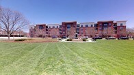 This multi-story residential building presents a classic and distinguished architectural design, constructed with rich red brick and accented with light-colored trim and large windows that line each floor. The exterior is well-proportioned, with multiple balconies offering residents private outdoor space and views of the surrounding grounds.

The building is set within a thoughtfully landscaped campus-style environment. A generous, well-manicured lawn stretches out in front of the structure, creating a sense of openness and providing ample green space. At the heart of the grounds, a central courtyard features a prominent statue as a focal point, surrounded by a circular path, mature trees, and park benches — offering a welcoming area for outdoor relaxation and community gathering.

The grounds are neatly maintained, with carefully laid pathways connecting parking areas to the building entrances. The surrounding trees, though seasonally bare in appearance, suggest a landscape that transforms with the seasons, providing shade and natural beauty throughout the year.

The overall ambiance of the property combines the warmth of classic brick architecture with the openness of its well-kept exterior spaces, creating an inviting and community-oriented environment under open skies.