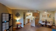 This single-floor unit offers a thoughtfully designed open-concept layout that maximizes the sense of space and flow between living areas. The kitchen is positioned at the heart of the home and features warm wood-tone cabinetry, a peninsula counter that doubles as a casual dining or prep surface, white appliances including a refrigerator, stove, and dishwasher, and a sink beneath a dedicated ventilation system. The peninsula creates a natural division between the kitchen and the adjacent dining area, which comfortably accommodates a full dining set. The living area extends seamlessly from the dining space, featuring rich dark hardwood-style flooring that runs consistently throughout the unit, lending a cohesive and polished aesthetic. Textured popcorn ceilings and neutral warm-toned walls provide a classic backdrop throughout. A hallway from the living area leads to additional rooms, with a visible closet offering built-in hanging storage. Central HVAC vents are positioned throughout the ceiling for efficient climate control. The overall ambiance is warm and functional, with a practical floor plan that makes excellent use of every square foot while maintaining an open and connected feel between all living spaces.