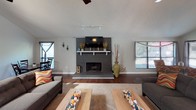 This single-story space makes a strong first impression with its dramatic vaulted ceilings that lend an expansive, airy quality throughout the main living areas. Rich, dark hardwood flooring flows seamlessly from room to room, providing a warm and cohesive foundation underfoot.

The living room is a standout, anchored by a painted dark brick fireplace with a white mantel that serves as a bold focal point. A mounted television sits above the mantel, flanked by windows dressed with blinds that allow for adjustable natural light. Dual ceiling fans with light fixtures enhance both comfort and style in this generous gathering space. A patio door provides access to the exterior, further brightening the room.

Adjacent to the living area, the dining space offers an intimate setting with recessed lighting and a framed wall mirror that reflects light and adds depth. The cool blue-grey accent wall adds a touch of color and sophistication. The open sightlines between the dining and living areas create a natural, connected flow ideal for entertaining.

A bar-height counter transitions into what appears to be a kitchen area, visible in the background with additional seating. Throughout the space, textured ceilings, crisp white trim, and neutral wall tones create a clean, inviting atmosphere that balances comfort with understated elegance.