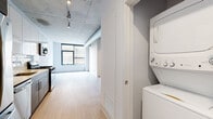 Located at 4555 N Sheridan Road in Chicago's vibrant Uptown neighborhood, this single-floor loft-style unit blends industrial character with modern finishes throughout. Exposed concrete ceilings and visible ductwork establish a distinctive urban aesthetic, while light wood flooring and crisp white walls keep the overall feel bright and contemporary.

The open living area is bathed in natural light from large industrial-style windows that frame views of the surrounding cityscape, creating an airy and spacious atmosphere. An exposed ventilation duct runs along the ceiling, adding to the loft's raw architectural charm.

The kitchen is outfitted with stainless steel appliances — including a gas range, built-in microwave, and refrigerator — complemented by two-tone cabinetry featuring white upper cabinets and gray lower cabinets, a white quartz countertop, and a classic subway tile backsplash. Light wood flooring flows seamlessly from the kitchen into the living space.

The bedroom offers a generous walk-in closet with built-in shelving, maintaining the unit's clean and functional design. The bathroom features a full soaking tub with a tiled surround, a modern floating vanity with a white countertop, large mirror, and gray tile flooring. A stacked washer and dryer are conveniently tucked into a dedicated in-unit laundry closet, completing this well-appointed urban residence.