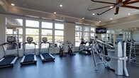 This fitness center is thoughtfully designed to provide a bright, functional workout environment. Large floor-to-ceiling windows line the walls, flooding the space with abundant natural light and offering views of the surrounding exterior. The open layout accommodates a variety of cardio equipment, including treadmills, stationary bikes, and elliptical machines, all arranged to maximize usability and comfort. Recessed lighting in the ceiling supplements natural light, ensuring the space remains well-lit at all times. A wall-mounted flat-screen television is positioned for easy viewing during workouts. The flooring features a durable dark surface well-suited for high-traffic fitness use. Crown molding along the upper walls adds a refined architectural detail, elevating the overall aesthetic beyond a typical gym setting. Mirrors are incorporated into the design, enhancing the sense of space and allowing users to monitor their form. The clean, neutral color palette of white and grey tones contributes to a modern, polished ambiance throughout the facility.