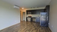 This single-floor space offers a clean, contemporary design centered around an open-concept living and kitchen area. The kitchen is well-appointed with dark espresso cabinetry that extends to the ceiling, providing ample storage. Stainless steel appliances — including a gas range with oven, built-in microwave, dishwasher, and a full-size refrigerator — lend a sleek, modern aesthetic. A track lighting system mounted above the kitchen ensures excellent task lighting throughout the cooking area. Rich, dark hardwood-style plank flooring runs throughout the main living space, adding warmth and visual depth to the open layout. A ductless mini-split air conditioning unit is mounted high on the wall, offering efficient and quiet climate control without the need for bulky ductwork. The neutral-toned walls and light ceiling create a bright, airy atmosphere that complements the darker accents of the cabinetry and flooring. A hallway visible from the main living area leads to additional rooms, suggesting a practical and well-organized floor plan. The overall ambiance is modern and functional, making this a highly livable and stylish space.