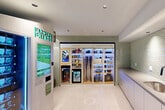This smartly designed amenity room offers a highly functional and modern space for building residents. The interior is defined by eye-catching sage green subway tile walls that lend a clean, contemporary aesthetic throughout. Large-format light stone floor tiles complement the bright, recessed-lit white ceiling, creating a crisp and welcoming atmosphere.

The centerpiece of the space is the Tulu Shop — a smart rental store offering an illuminated display of everyday essentials and rentable items such as vacuums, projectors, snacks, and beverages, all accessible via an interactive kiosk. Adjacent to it, a Farmers Fridge automated machine provides fresh salads, bowls, and sandwiches, making healthy food conveniently available around the clock.

Along one wall, a continuous run of sleek gray cabinetry is topped with a smooth countertop and fitted with a stainless steel sink, offering a practical utility station. Black-framed glass double doors provide access to the broader building corridor, while a large interior window maintains visual connectivity with adjacent spaces. The overall design balances utility and aesthetics, delivering a well-equipped, technology-forward amenity that serves the everyday needs of its users with efficiency and style.