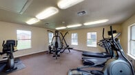 This fitness center is a dedicated, single-floor workout facility designed to support a range of exercise routines. The space is generously sized, offering ample room for users to move freely between equipment without feeling crowded. The layout is practical and functional, with carpeted flooring and protective rubber mats placed beneath the machines for added safety and durability.

The facility is equipped with a comprehensive selection of fitness equipment, including multiple treadmills with digital displays, elliptical trainers, a multi-station cable weight machine, a rowing machine, and additional strength training equipment. A wall-mounted television is positioned near the treadmill area, allowing users to stay entertained during cardio sessions.

Natural light filters into the space through numerous windows along the walls, offering views of the surrounding outdoor greenery. The bright, neutral-toned walls contribute to an open and energizing atmosphere. Overhead fluorescent lighting ensures the space remains well-lit throughout the day.

A dedicated entry door provides direct exterior access, and the facility also includes practical amenities such as a wall-mounted exercise chart, a fire extinguisher, and a thermostat for climate control. The clean, organized layout makes this fitness center a convenient and accessible option for a variety of fitness levels and goals.