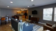 This single-floor space showcases a well-designed open-concept layout that connects the living area and kitchen in one cohesive, functional environment. The living area is anchored by a comfortable sectional sofa in cool blue-grey tones, complemented by a striped area rug and a sleek metal-framed coffee table with a woven centerpiece. A wall-mounted flat-screen television sits above a mid-century modern style credenza in warm walnut tones, adding both style and storage. Recessed lighting throughout the ceiling provides even, warm illumination across the space, while a window fitted with white shutters allows natural light to filter in softly.

The kitchen flows seamlessly from the living area and features warm wood cabinetry, black appliances, and a peninsula countertop with bar seating — ideal for casual dining or entertaining. Pendant lights hang above the kitchen area, adding a decorative touch that enhances the warm, inviting atmosphere. The flooring transitions smoothly between tile and wood-look surfaces, tying the two areas together cohesively.

Overall, the space presents a modern yet comfortable aesthetic, defined by its dark grey accent walls, warm wood tones, and thoughtfully chosen furnishings that create a balanced and livable environment.