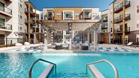 Located at 2929 North Mayfair Road in Wauwatosa, Wisconsin, this contemporary multi-story residential community is defined by its striking exterior architecture, blending wood-accented facades, dark brick detailing, and private balconies that overlook a thoughtfully designed outdoor amenity complex. The heart of the property is a sprawling courtyard that serves as a social and recreational hub for residents. A resort-style swimming pool takes center stage, complete with an elegant pergola structure featuring cascading water curtains, complemented by an expansive sun deck lined with lounge chairs for relaxing under open skies. Adjacent to the pool area, a dedicated fire pit lounge furnished with vibrant yellow Adirondack chairs and plush seating creates an inviting atmosphere for evening gatherings. A regulation bocce ball court and a well-manicured turf lawn offer additional outdoor recreation, while decorative planters with seasonal blooms add color and warmth throughout the courtyard. Cornhole and casual seating areas round out the recreational offerings. The entire outdoor space is enclosed by a gated perimeter, ensuring privacy and security. The building's multi-story design, with numerous private balconies framing the courtyard, creates a welcoming, community-centered environment bathed in natural light.