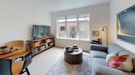 This thoughtfully designed single-floor residence blends modern aesthetics with functional living. The open-concept layout connects the living area and kitchen, creating a cohesive and comfortable space. The living room features large windows that invite abundant natural light and offer views of the surrounding brick-clad neighborhood. Light hardwood-style flooring runs throughout, complementing the clean white walls and contemporary finishes. A cozy seating area anchors the living space, accented by bold artwork and warm lighting.

The kitchen is equipped with stainless steel appliances, including a range, built-in microwave, and dishwasher. Two-tone cabinetry — dark lower cabinets paired with crisp white upper cabinets — frames a classic white subway tile backsplash and light-toned countertops, giving the kitchen a clean, modern look.

The bedroom is a calm retreat, featuring light wood flooring, a spacious walk-in closet with open shelving, and a frosted glass entry door that adds a sleek, modern touch. Wall-mounted lighting and ample storage contribute to the room's understated elegance.

The bathroom is finished in a minimalist style with light wood-tone flooring, white walls, a dark wood vanity, and clean fixtures, creating a serene and practical space. Overall, the residence presents a cohesive modern design with attention to detail throughout.