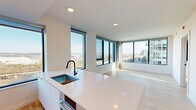 Located on the seventh floor at 3130 Potomac Ave in Arlington, VA, this contemporary 2-bedroom, 2-bathroom residence offers a thoughtfully designed open layout with abundant natural light throughout. The combined kitchen and living area serves as the heart of the home, featuring sleek white flat-front cabinetry that extends to the ceiling, stainless steel appliances including a gas range and wall oven, a stainless steel range hood, and a large white quartz island with an undermount sink and matte black fixtures. Expansive floor-to-ceiling windows wrap around the living space, framing panoramic views of the Arlington cityscape and beyond. Light-toned hardwood floors run throughout the living areas and both bedrooms, lending warmth and continuity to the modern aesthetic. The primary bedroom is generously sized, offering wide corner windows with sweeping views of the surrounding urban landscape. The second bedroom also benefits from large picture windows with bright, open outlooks. Both bathrooms are finished with a clean, contemporary palette, featuring wide-format tile flooring, white cabinetry with warm stone-toned countertops, undermount sinks, matte black hardware, and backlit LED vanity mirrors. The primary bathroom includes a dual-sink vanity, adding functionality to its refined design. A hallway and dedicated closet provide additional storage and circulation throughout the residence.