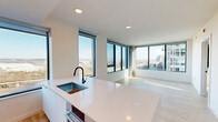 Located on the seventh floor at 3130 Potomac Ave in Arlington, VA, this contemporary 2-bedroom, 2-bathroom residence offers a thoughtfully designed open layout with abundant natural light throughout. The combined kitchen and living area serves as the heart of the home, featuring sleek white flat-front cabinetry that extends to the ceiling, stainless steel appliances including a gas range and wall oven, a stainless steel range hood, and a large white quartz island with an undermount sink and matte black fixtures. Expansive floor-to-ceiling windows wrap around the living space, framing panoramic views of the Arlington cityscape and beyond. Light-toned hardwood floors run throughout the living areas and both bedrooms, lending warmth and continuity to the modern aesthetic. The primary bedroom is generously sized, offering wide corner windows with sweeping views of the surrounding urban landscape. The second bedroom also benefits from large picture windows with bright, open outlooks. Both bathrooms are finished with a clean, contemporary palette, featuring wide-format tile flooring, white cabinetry with warm stone-toned countertops, undermount sinks, matte black hardware, and backlit LED vanity mirrors. The primary bathroom includes a dual-sink vanity, adding functionality to its refined design. A hallway and dedicated closet provide additional storage and circulation throughout the residence.