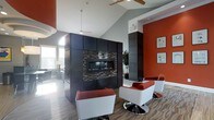 This single-floor space makes a strong design statement through its bold use of color, materials, and architectural detail. The centerpiece of the main lounge area is a sleek, built-in linear fireplace framed by a striking floor-to-ceiling surround clad in dark cabinetry and mosaic tile accents, creating a dramatic visual anchor for the room. Soaring vaulted ceilings amplify the sense of openness, while ceiling fans provide both function and a clean, modern aesthetic overhead. The space is brought to life by a vivid color scheme — deep burnt orange accent walls contrast sharply against cool grey tones throughout — lending the interior an energetic yet sophisticated character. Contemporary white and red swivel chairs are arranged around the fireplace, offering a stylish and inviting seating area. Hardwood-style flooring and patterned area rugs add warmth and texture underfoot. Natural light filters in through large windows, brightening the space and complementing the thoughtfully layered artificial lighting, including recessed and track fixtures. Lush indoor greenery adds a refreshing natural element, softening the boldness of the overall design. The layout flows openly, making the space feel both functional and visually dynamic.