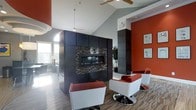 This single-floor space makes a strong design statement through its bold use of color, materials, and architectural detail. The centerpiece of the main lounge area is a sleek, built-in linear fireplace framed by a striking floor-to-ceiling surround clad in dark cabinetry and mosaic tile accents, creating a dramatic visual anchor for the room. Soaring vaulted ceilings amplify the sense of openness, while ceiling fans provide both function and a clean, modern aesthetic overhead. The space is brought to life by a vivid color scheme — deep burnt orange accent walls contrast sharply against cool grey tones throughout — lending the interior an energetic yet sophisticated character. Contemporary white and red swivel chairs are arranged around the fireplace, offering a stylish and inviting seating area. Hardwood-style flooring and patterned area rugs add warmth and texture underfoot. Natural light filters in through large windows, brightening the space and complementing the thoughtfully layered artificial lighting, including recessed and track fixtures. Lush indoor greenery adds a refreshing natural element, softening the boldness of the overall design. The layout flows openly, making the space feel both functional and visually dynamic.