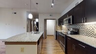 This well-designed single-floor residence showcases a cohesive modern aesthetic throughout its layout. The kitchen serves as the centerpiece of the unit, offering a striking combination of dark espresso cabinetry reaching floor to ceiling, light granite countertops, and a full suite of stainless steel appliances including a refrigerator, range, and built-in microwave. A mosaic tile backsplash adds texture and visual interest, while warm hardwood-style flooring runs throughout. A central island with an integrated sink and pendant lighting above provides both functional workspace and a natural gathering point. Track lighting further illuminates the space efficiently. The bathroom continues the design language with matching dark cabinetry, granite vanity top, a framed mirror, and a tub-shower combination. Ample storage is provided by a tall linen cabinet alongside the vanity. The bedroom is a comfortable, quieter retreat featuring plush carpeting, a ceiling fan, and multiple windows dressed with slatted blinds that invite generous natural light. Clean white walls and consistent trim work tie the entire unit together, creating a polished and functional living environment.
