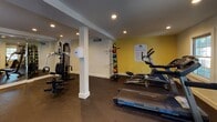 Located at 1244 Hamilton Ct in Cary, NC, this single-level fitness center is part of the Williamsburg community and offers a comprehensive workout facility designed to meet a variety of exercise needs. The space is organized into distinct zones, seamlessly connected and thoughtfully laid out for functionality and flow.

The cardio area features multiple Nautilus treadmills positioned in front of a wall-mounted flat-screen television, providing entertainment during workouts. Natural light filters in through windows, complementing the recessed ceiling lighting that illuminates the entire facility. Rubber flooring throughout ensures safety and durability.

A dedicated strength training section houses a multi-function cable machine and free weight accessories, including medicine balls and stability balls neatly stored on racks. Mirrored walls in this area enhance the sense of openness and allow users to monitor their form. A bright yellow accent wall adds a bold, energizing touch to the space.

A standout architectural feature is the floor-to-ceiling glass partition wall etched with the Williamsburg logo, which separates the fitness floor from an adjacent glass-enclosed studio or multipurpose room. This transparent divider maintains visual connectivity while defining separate functional areas. The overall design is clean, modern, and well-maintained, reflecting a community-focused amenity space built for everyday use.