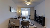 This single-floor residence offers a thoughtfully designed layout with interconnected living spaces that create an open, welcoming atmosphere. The main living area is anchored by light wood-style flooring, a ceiling fan with integrated lighting, and neutral-toned walls that provide a versatile backdrop for any décor style. A plush area rug with a bold geometric pattern adds texture and visual interest to the space, complemented by comfortable seating arrangements including a large sofa and accent chairs in warm, earthy tones. A sleek media console along one wall provides both function and style. An arched passageway leads seamlessly into a secondary sitting or bonus room, which is bathed in natural light streaming through a large multi-pane window overlooking the outdoors. This adjoining space features its own ceiling fan and offers a bright, airy retreat ideal for reading or relaxing. The consistent flooring and color palette throughout both areas create a cohesive flow. Wall art and floor lamps add decorative touches that enhance the overall ambiance, making this residence feel both elegant and livable.