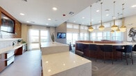 This thoughtfully designed single-floor amenity lounge combines contemporary style with warm, eclectic details to create a dynamic and welcoming communal space. The layout flows across several distinct zones, each serving a different social function while maintaining a cohesive aesthetic throughout.

At the heart of the space is a striking bar area anchored by a bold herringbone-patterned tile accent wall in rich amber and teal tones, complemented by a white subway tile backsplash, granite countertops, stainless steel under-counter refrigerators, and bar seating with sleek black chairs. Gold globe pendant lights hang above, adding warmth and visual interest.

Adjacent to the bar, a curved teal velvet banquette encased in warm wood paneling offers an intimate seating nook, while nearby café-style tables and chairs provide flexible gathering options. A wall-mounted television in this zone makes it ideal for casual viewing or socializing.

A separate lounge area features a plush navy sectional sofa, tan leather accent chairs, and a round coffee table, all arranged around natural window light and a dramatic reclaimed-wood television feature wall. The space is bright and comfortable, with views of the surrounding cityscape visible through large windows throughout.
