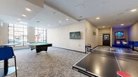 Located at 70 Park East Drive in Winnipeg, this single-floor amenity games room offers a generously sized recreational space designed for socializing and active play. The layout flows through several interconnected zones, all finished with neutral-toned patterned carpet, dark baseboards, and smooth white walls illuminated by recessed pot lighting throughout.

The main area is anchored by a full-size billiards table positioned near large floor-to-ceiling windows that flood the space with natural light and provide views to the exterior streetscape. A wall-mounted flat-screen television is centrally placed for easy viewing. Brunswick ping pong tables occupy their own dedicated zone, offering ample room for competitive play.

A secondary alcove features a tall bistro-style table with bright blue chairs alongside an arcade basketball shooting game, creating a casual lounge nook adjacent to the clearly marked Fitness Centre entrance. A separate, quieter room houses a full-length shuffleboard table, providing a more relaxed gaming option.

The overall ambiance is modern and activity-focused, with a clean, well-maintained aesthetic suited to a residential amenity setting. The thoughtful zoning of the space allows multiple activities to take place simultaneously, making it a versatile hub for recreation and community engagement.