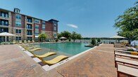 This expansive outdoor amenity area is centered around a generously sized resort-style swimming pool, designed to offer both relaxation and striking visual appeal. The pool deck is finished in a distinctive herringbone-pattern brick, lending a warm, upscale character to the entire space. In-pool lounge chairs in bold yellow invite residents to enjoy the shallow wading areas, while traditional chaise lounges and shaded umbrella seating are arranged along the pool perimeter for additional comfort.

The space is framed by a multi-story residential building constructed with a blend of rich brick and natural stone masonry, giving the complex a refined Hill Country-inspired aesthetic. A prominent stone archway serves as a key architectural focal point at the pool entrance, complemented by large glass panels and teal standing-seam metal roof accents that echo throughout the community's design.

Lush, drought-tolerant landscaping featuring ornamental grasses, agave, and native plantings lines the walkways and borders, adding texture and greenery to the hardscaped surfaces. At the far end of the pool, an open waterfront fence preserves unobstructed views of a tranquil lake, with mature trees framing the horizon. The overall ambiance is that of a polished, resort-caliber outdoor retreat bathed in abundant natural sunlight.