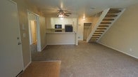 This two-story space is designed with an open-concept layout on the main floor, seamlessly connecting the living area and kitchen. The living space features neutral-toned carpet that extends throughout, complemented by a parquet wood floor entry that adds a classic touch near the front. A ceiling fan with a light fixture anchors the living area, providing both comfort and illumination.

The kitchen is positioned at the heart of the main floor, defined by a raised breakfast bar that serves as a natural divider between the cooking and living spaces. White cabinetry lines the upper portion of the kitchen, offering ample storage, while black appliances — including a microwave and refrigerator — provide a clean contrast. Under-cabinet lighting adds warmth to the workspace.

A straight staircase with open wood-tread steps and a white railing leads to the second floor, contributing to the open and airy feel of the space. Beneath the staircase, a storage closet with shelving offers practical organizational space. The neutral color palette and warm lighting throughout create a welcoming and functional atmosphere across both levels.