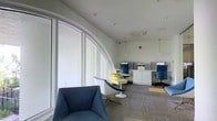 This single-floor space offers a well-designed, functional environment that blends modern aesthetics with practical workspace amenities. A striking architectural feature is the curved, floor-to-ceiling glass window wall along one side of the room, allowing abundant natural light to fill the interior and creating an open, airy atmosphere. The white walls and light ceiling complement the brightness introduced by this generous glazing.

The space is divided into distinct functional zones. A lounge area near the windows features contemporary seating, including a navy blue upholstered armchair and a sleek white pod-style chair paired with a small round side table, offering a comfortable setting for informal meetings or relaxation. Further into the space, a dedicated workstation area includes multiple computer desks equipped with monitors, separated by low privacy dividers, and fitted with ergonomic blue task chairs. White storage cabinets beneath the workstations provide practical organizational solutions.

Pendant lighting hangs from the ceiling alongside recessed spotlights and a centrally mounted HVAC unit, ensuring the space remains well-lit and climate-controlled. The patterned carpet tile flooring with yellow accent squares ties the color scheme together, referencing the yellow accents found in the seating, contributing to a cohesive and professional overall ambiance.