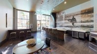 Located along Fletcher Parkway in La Mesa, California, this single-story space presents a thoughtfully designed interior that blends modern aesthetics with a coastal-inspired atmosphere. The open layout is anchored by soaring ceilings with a distinctive gold-toned accent, creating an airy and energetic ambiance throughout.

A standout feature is the dramatic floor-to-ceiling black-and-white photographic mural depicting a surfer against a coastal cityscape, lending the space a bold, urban-beach character. Directly in front of the mural sits a long, curved bar counter with a light-colored surface and rich wood base, accompanied by a row of modern bar-height seating and pendant lighting that hangs overhead.

The main gathering area includes a round table surrounded by sleek black leather chairs, set atop a vibrant, multicolored area rug that adds warmth and contrast to the dark hardwood-style flooring. A wall-mounted flat-screen TV is positioned to serve this seating zone. Oversized windows line the walls, flooding the interior with natural light and framing lush greenery outside. A glass block partition wall adds textural interest and allows diffused light to filter through, enhancing the overall brightness and openness of the space.