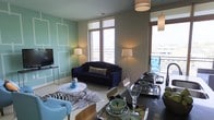 This single-floor space showcases a thoughtfully designed open-concept layout that seamlessly connects the living and dining areas. The living room is a standout, featuring an accent wall painted in a soft mint green adorned with white geometric panel molding, adding a layer of sophistication and visual interest. A deep navy blue sofa serves as the focal point, complemented by eclectic décor including a zebra-print area rug and a round white coffee table.

Large floor-to-ceiling windows flood the interior with generous natural light while offering views of the surrounding cityscape. The windows open to a balcony, extending the living space outdoors and enhancing the airy, open ambiance.

The dining area sits adjacent to the living room and features a sleek dark dining table set with stylish glassware and decorative bowls, lit by contemporary pendant lighting overhead. Warm-toned walls in the dining zone create a subtle contrast to the mint green living area, adding depth and dimension to the overall aesthetic.

Throughout the space, the interplay of bold colors, layered textures, and modern furnishings creates a dynamic yet cohesive environment that feels both functional and design-forward.