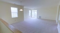 This single-floor space offers a clean, open layout designed to maximize comfort and functionality. The main living area is generously sized, featuring plush wall-to-wall carpeting that adds warmth underfoot, while smooth hardwood flooring defines the adjacent dining or entryway zone, creating a natural visual distinction between spaces.

Natural light is a defining characteristic throughout, with multiple large windows dressed in white trim that invite abundant daylight and offer views of the surrounding greenery. A set of glass doors leads directly to an outdoor balcony, further enhancing the sense of openness and providing a seamless connection to the outdoors.

The interior palette is kept neutral, with soft cream-toned walls and white trim that lend a fresh, versatile canvas well-suited to a variety of furnishing styles. A single pendant light fixture hangs centrally, providing focused illumination while complementing the understated aesthetic of the space.

HVAC venting is integrated discreetly into the walls and floor, ensuring climate comfort year-round. The overall ambiance is light, airy, and move-in ready, offering a functional and pleasant living environment with thoughtful design details throughout.