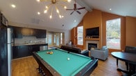 This single-story clubhouse is thoughtfully designed to serve as a social hub, combining entertainment, comfort, and functionality under one roof. The space is anchored by dramatic vaulted ceilings with exposed beams, which lend an open, airy feel throughout. A striking sputnik chandelier and ceiling fan hang overhead, complementing the warm wood-look flooring that extends across much of the main area.

The centerpiece of the entertainment zone is a full-size pool table positioned beneath the statement lighting. Adjacent to it, a comfortable lounge area features leather sofas and accent chairs arranged around a tile-surround gas fireplace. A wall-mounted flat-screen TV with built-in shelving sits above the fireplace, creating an ideal media viewing setup. Natural light filters in through multiple windows, with views hinting at an outdoor pool area nearby.

The entry foyer transitions smoothly from tile flooring into the main space, accented with decorative consoles, framed artwork, and colorful wall pieces that add a welcoming character. A compact kitchenette at the rear is outfitted with dark cabinetry, granite-style countertops, a stainless steel refrigerator, microwave, and coffee station, making it well-equipped for hosting events.