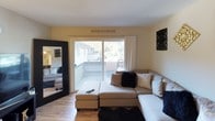 Located at 7051 Bowling Dr in Sacramento, California, this single-floor unit offers a thoughtfully arranged layout that maximizes comfort and functionality. The living room serves as the heart of the space, featuring warm hardwood-style flooring, neutral walls, and a large sliding glass door that opens to a private balcony with tree-lined views, filling the room with natural light. Decorative wall art in black and gold tones adds a modern, curated aesthetic throughout.

The kitchen is efficiently designed with white cabinetry, white appliances including a full-size refrigerator, and light tile flooring. A side window provides natural light, and a corner shelving unit adds both storage and a decorative touch.

The bedroom offers a calm and restful atmosphere with plush carpeting and natural light from a window. A dedicated vanity area with a lighted mirror provides a functional grooming space, making the room both practical and comfortable.

The bathroom features a granite-topped vanity with a single sink and clean, light-toned finishes. A hallway connects the rooms seamlessly, with hardwood-style flooring continuing the cohesive design of the space. Overall, the unit presents a well-maintained and stylishly decorated interior with a consistent black, white, and gold color palette.
