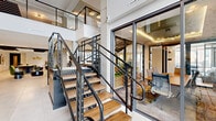 This generously proportioned amenity space showcases a sophisticated modern design across a sprawling open floor plan. The multi-level layout is anchored by a striking open-tread staircase with sleek steel cable railings and warm wood steps, connecting mezzanine areas enclosed by glass panel railings that allow light and sightlines to flow freely throughout.

The living area centers around a stunning floor-to-ceiling marble slab feature wall housing a wide linear fireplace, flanked by open wood shelving and complemented by plush green velvet seating. A cozy library nook, finished in deep navy with built-in bookshelves, wall sconces, and an area rug, offers a more intimate retreat within the larger space.

The dining area accommodates generous group gatherings around a dark wood table set with bold yellow upholstered chairs, bathed in natural light from large windows and sliding glass doors. A well-appointed kitchen corridor features under-counter refrigeration units, open display shelving, and pendant lighting. A charming banquette nook, accented in green with plaid upholstery and lush tropical plants, adds a casual dining option.

Large-format porcelain tile flooring flows throughout, while curated artwork, neon signage, and warm metallic accents lend the space a vibrant yet refined ambiance.