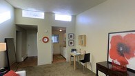 This single-floor space is thoughtfully laid out to maximize functionality and comfort. The main living area features neutral carpet flooring and is bathed in natural light streaming through a row of clerestory windows positioned high along the upper wall, creating a bright and airy atmosphere without compromising wall space or privacy. The textured ceiling adds subtle visual character throughout.

An open passageway connects the living area to the kitchen, which features hardwood-style flooring, white cabinetry, white appliances including a refrigerator, and a window that allows additional natural light into the space. The kitchen layout offers practical counter space and storage, well-suited for everyday use.

Adjacent to the living area, a dedicated dining nook accommodates a small table and seating, making efficient use of the available space. A wall-mounted organizer and framed artwork add personality and warmth to the room.

A separate room visible from the living area includes a spacious closet, suggesting a well-defined bedroom area with ample storage. The overall color palette of soft whites and warm neutrals throughout the space creates a cohesive, calm, and welcoming ambiance ideal for comfortable day-to-day living.