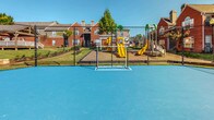 This outdoor recreational area is situated within a brick residential community and offers a variety of amenities for active use. The centerpiece is a spacious, bright blue sports court with clearly marked lines and a goal post, suitable for multiple athletic activities. The smooth, hard surface provides ample room for play and is fully enclosed by a chain-link fence, ensuring a defined and secure boundary for the court.

Adjacent to the sports court is a dedicated children's playground equipped with colorful play structures, including yellow and green slides and climbing elements, all set on a mulched surface for safety. Seating benches are positioned nearby, offering a comfortable vantage point for spectators or those supervising children.

The surrounding grounds are well-maintained, featuring manicured lawns, mature trees, and landscaped shrubs that contribute to the area's overall greenery. Concrete pathways and steps connect the recreational space to the surrounding buildings, which are constructed of classic red brick with gray metal railings and staircases. The open-air setting benefits from abundant natural light and clear sky views, creating an inviting and energetic outdoor environment ideal for community recreation.