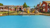 This outdoor recreational area is situated within a brick residential community and offers a variety of amenities for active use. The centerpiece is a spacious, bright blue sports court with clearly marked lines and a goal post, suitable for multiple athletic activities. The smooth, hard surface provides ample room for play and is fully enclosed by a chain-link fence, ensuring a defined and secure boundary for the court.

Adjacent to the sports court is a dedicated children's playground equipped with colorful play structures, including yellow and green slides and climbing elements, all set on a mulched surface for safety. Seating benches are positioned nearby, offering a comfortable vantage point for spectators or those supervising children.

The surrounding grounds are well-maintained, featuring manicured lawns, mature trees, and landscaped shrubs that contribute to the area's overall greenery. Concrete pathways and steps connect the recreational space to the surrounding buildings, which are constructed of classic red brick with gray metal railings and staircases. The open-air setting benefits from abundant natural light and clear sky views, creating an inviting and energetic outdoor environment ideal for community recreation.