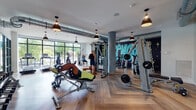 Located at 1661 NW 16th St in Miami, FL, this single-level fitness center offers a well-appointed and energetic environment designed for a comprehensive workout experience. The space is divided into two primary zones: a main strength and cardio training area and a separate group fitness studio, both unified by a distinctive herringbone-patterned wood floor that lends a contemporary, upscale feel throughout.

The main training area is equipped with a range of cable machines, weight benches, and free weights, complemented by a row of treadmills and cardio machines positioned along floor-to-ceiling windows. These expansive windows flood the space with natural light and provide views of the surrounding greenery, creating an open and inviting atmosphere. Exposed ductwork runs along the white ceiling, while pendant lights with matte black shades add an industrial-modern touch. A bold, colorful graffiti mural anchors one wall, reinforcing the facility's urban, energetic character.

The adjoining studio is enclosed by full glass walls and features floor-to-ceiling mirrors on two sides, a large mounted flat-screen display, spin bikes, stability balls, and a rowing machine. Black-framed windows continue the modern aesthetic while maintaining visual connectivity to the outdoors. The overall ambiance is dynamic, bright, and motivating.