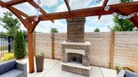 This outdoor space centers around a striking stacked stone fireplace that serves as the focal point of a well-designed patio area. The fireplace is constructed with layered natural stone in warm gray and tan tones, accented by a smooth light-colored mantel and hearth surround that adds a refined contrast to the textured stonework. The chimney rises prominently, giving the structure a substantial, architectural presence.

Overhead, a sturdy wood pergola with rich warm-toned beams provides partial overhead coverage, with an open-sky design that allows natural light to filter through. Decorative globe string lights are strung beneath the pergola, adding ambiance for evening gatherings. The concrete patio floor offers a clean, durable surface underfoot.

Privacy is well-established by a horizontal-plank cedar fence that encloses the space on multiple sides, creating an intimate and secluded atmosphere. A tall square planter with seasonal greenery adds a natural decorative touch near the fireplace.

Overall, this outdoor living area is thoughtfully designed for year-round entertaining, combining warmth, privacy, and style in a functional and inviting setting.