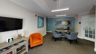 Located at 25 Landis in Lillington, NC, this single-floor professional office space is thoughtfully designed to balance functionality with a welcoming atmosphere. The interior is finished with neutral walls accented by a distinctive teal feature wall, crown molding, and textured ceilings that lend the space a polished, professional character throughout.

The main area serves as both a reception and consultation zone, centered around a large work desk accompanied by a set of matching gray upholstered chairs arranged for client-facing interactions. A second workstation with built-in cabinetry sits against the teal accent wall, offering an organized and efficient workspace. Recessed and overhead fluorescent lighting provides consistent, well-distributed illumination across the entire floor.

A comfortable waiting area is integrated into the space, featuring a bold orange tufted accent chair and a media console with a wall-mounted flat-screen television, creating an inviting environment for visitors. A vibrant abstract painting in teal and gold tones adds a dynamic focal point to this area. Additional seating is available near the entrance, where a pair of chairs flank a set of glass-paneled double doors that fill the entry with natural light. Carpet flooring runs throughout, contributing to a quiet and professional ambiance. The space also includes a framed celestial triptych artwork that adds a distinctive artistic touch to the overall design.