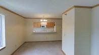 This single-floor residence offers a clean, open-concept layout designed for comfortable everyday living. The living area is finished with neutral carpeting and light-toned walls accented by warm wood crown molding and baseboards, creating a cohesive and inviting atmosphere throughout.

A defining feature of the space is the pass-through opening between the living and kitchen areas, which promotes an open flow and allows for easy interaction between rooms. The kitchen is equipped with warm honey-toned wood cabinetry providing ample storage, a white microwave, and a refrigerator. The pass-through ledge doubles as a convenient breakfast bar or serving counter, adding to the practicality of the layout.

Overhead, a classic chandelier with multiple lights serves as a focal point in the dining area, casting warm light across the space. The textured ceiling adds subtle visual interest, while the consistent wood trim detailing throughout ties the design together with a traditional touch.

The layout is straightforward and efficient, making excellent use of the available floor space. The neutral color palette and clean finishes give the space a bright, move-in-ready feel, well-suited for a variety of furnishing styles and personal tastes.