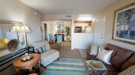 This single-floor residence offers a thoughtfully arranged open-concept layout that seamlessly connects the living, dining, and kitchen areas, creating a cohesive and functional flow throughout the space. The living area is warm and inviting, featuring neutral-toned walls and carpeted flooring accented by tiled entryways, providing both comfort and practicality. The space is furnished to maximize comfort, with plush seating and cheerful aqua and teal accents that lend a light, coastal-inspired ambiance. A striped area rug anchors the living area and adds a touch of color and texture to the room. Beyond the living space, the dining area provides an intimate setting for meals, accented by a warm lamp and a tall decorative plant that adds a natural, refreshing element. The kitchen is partially open to the main living area via a half-wall pass-through, featuring light cabinetry, standard appliances including a microwave and refrigerator, and a practical layout suited for everyday meal preparation. Natural light filters into the space, contributing to the overall bright and airy feel. The unit also includes an entryway with a front door, and additional rooms are accessible through interior doorways, suggesting private bedroom and bathroom accommodations. Overall, the space presents a comfortable, well-organized home with a cohesive and cheerful design aesthetic.