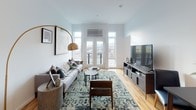 This single-floor residence showcases a thoughtfully designed layout with warm light wood flooring flowing throughout. The living area is a standout space, anchored by French doors that open to an outdoor area and frame sweeping views of the surrounding cityscape. Generous wall space accommodates curated artwork, while a wall-mounted climate control unit ensures year-round comfort. The modern kitchen is outfitted with dark cabinetry, stainless steel appliances including a French door refrigerator, electric range, dishwasher, and microwave, complemented by a stainless steel farmhouse sink, white subway tile backsplash, and open wood shelving for functional display. The bedroom offers a serene retreat, accessed through a sliding barn door, with a window framing outdoor views and ample natural light. The full bathroom features a soaking tub with shower, a dark-cabineted vanity with a white countertop, large mirror, and light wood flooring accented by a charcoal bath mat. Throughout the space, crisp white walls provide a clean, contemporary backdrop that enhances the overall bright and welcoming ambiance.