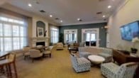 This single-floor communal space exudes a refined, classic elegance designed for comfort and socialization. The expansive lounge area is anchored by a stately white fireplace framed by a traditional mantelpiece, adding a warm focal point to the room. Arched architectural niches along the walls lend a sophisticated design element, complementing the rich teal and cream color palette that runs throughout the space.

Recessed lighting illuminates the room from above, highlighting the well-maintained neutral carpeted flooring beneath. The layout accommodates multiple distinct seating clusters, furnished with a tasteful mix of upholstered armchairs, sofas, and accent tables — offering flexible arrangements suited to both intimate conversation and larger gatherings.

Large windows dressed with plantation shutters line the walls, allowing abundant natural light to fill the interior while maintaining privacy. Doorways and pass-throughs connect to adjacent rooms, hinting at additional spaces beyond. Lush indoor plants are thoughtfully placed throughout, adding a touch of greenery and life to the environment.

Overall, the space presents a polished, welcoming atmosphere characteristic of a well-appointed community or hospitality setting, balancing aesthetic appeal with practical comfort.
