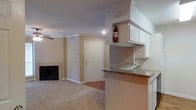 This single-floor residence offers a well-designed open-concept layout that seamlessly connects the kitchen and living areas. The kitchen is equipped with white upper cabinetry, dark granite countertops, and a peninsula with a sink that overlooks the main living space, making it ideal for both everyday functionality and entertaining. The living area features plush carpeting, a classic wood-burning fireplace with a tiled surround, and a ceiling fan, creating a warm and comfortable atmosphere. Crown molding runs throughout the space, adding a touch of refined elegance to the neutral, light beige walls. The entryway is framed by a paneled front door, providing a clean and welcoming transition into the home. Recessed lighting and overhead fixtures ensure the space remains well-lit throughout. The combination of hard flooring near the entry and soft carpet in the living area adds textural variety and practicality. Overall, the space presents a cozy yet functional living environment with thoughtful architectural details that elevate its everyday appeal.