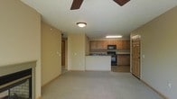 This single-floor residence offers a thoughtfully designed open-concept layout that seamlessly connects the living area and kitchen, creating a spacious and functional environment. The living space is finished with neutral-toned carpeting and warm beige walls, lending a cozy and welcoming ambiance throughout. A ceiling fan is centrally positioned overhead, providing both comfort and a casual aesthetic touch. The kitchen is defined by a peninsula-style counter that serves as a natural divider between the cooking and living areas. Warm wood-toned cabinetry lines the kitchen walls, offering generous storage, while dark granite-style countertops provide a contrasting accent. Black appliances, including a range and an over-the-range microwave, complete the kitchen setup. Recessed and flush-mount lighting ensures the space is well-lit throughout. A hallway visible from the main living area leads to additional rooms, suggesting a practical and well-organized floor plan. The overall design emphasizes ease of living, with clean lines, neutral finishes, and an efficient use of space that suits a variety of lifestyles.