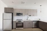 Unit 506 at 1213–1221 Broad St offers a thoughtfully designed one-bedroom, one-bathroom layout with a clean, modern aesthetic. Light hardwood flooring runs throughout the living areas and bedroom, creating a bright and cohesive feel across the entire floor plan.

The kitchen is a standout feature, boasting an L-shaped configuration with a peninsula that opens to the living area. Taupe wood-grain cabinetry extends to the upper walls, complemented by a white subway tile backsplash and light quartz countertops. Stainless steel appliances — including a refrigerator, electric range, microwave, and dishwasher — complete the space, while pendant lighting above the peninsula adds a modern finishing touch.

The bedroom is a well-proportioned retreat featuring a large window that draws in natural light and provides outdoor views. A wall-mounted climate control unit ensures year-round comfort, and a set of double-door closets offers generous storage capacity.

The bathroom features neutral tile flooring, a dark-toned vanity, and matte black hardware and grab bar accents, lending a contemporary contrast to the clean white walls. Panel-style doors with matte black hardware are found throughout the unit, adding a consistent design detail that ties the space together seamlessly.